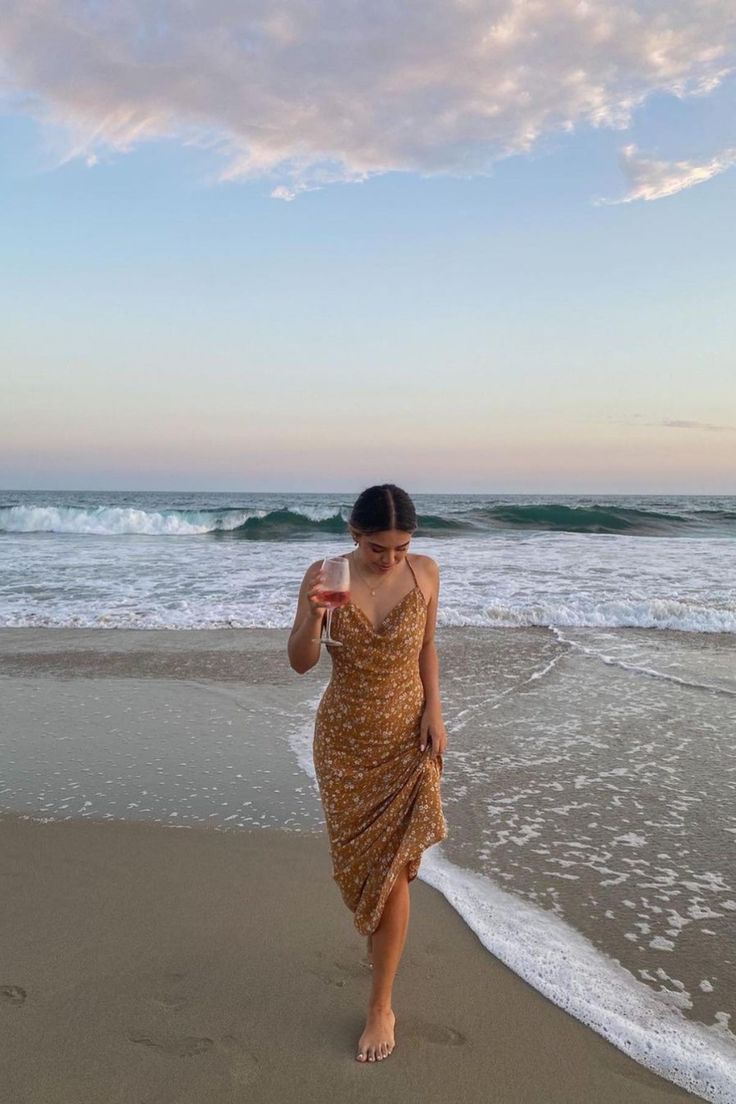 a woman walking on the beach holding a drink in her hand and looking at the ocean