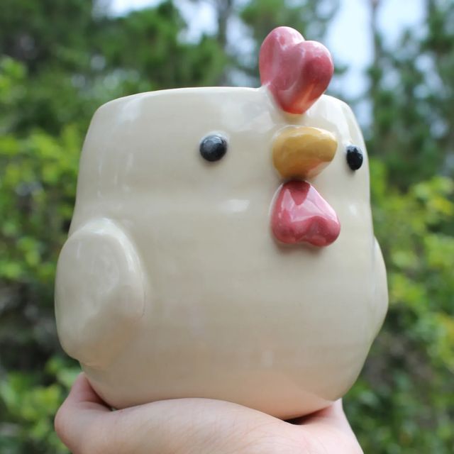 a hand holding a white ceramic chicken figurine in front of trees and bushes