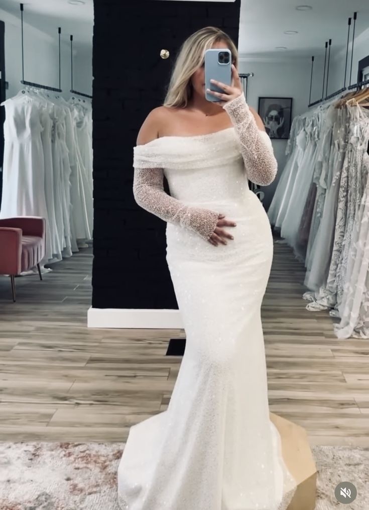 a woman taking a selfie with her phone in a wedding dress shop wearing a white gown