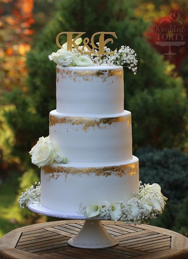 a white wedding cake with gold lettering and flowers on top is sitting on a wooden table