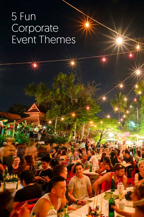 a large group of people sitting at tables in front of trees with lights on them