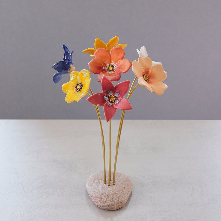 four colorful flowers in a vase on a white table top with grey wall behind them