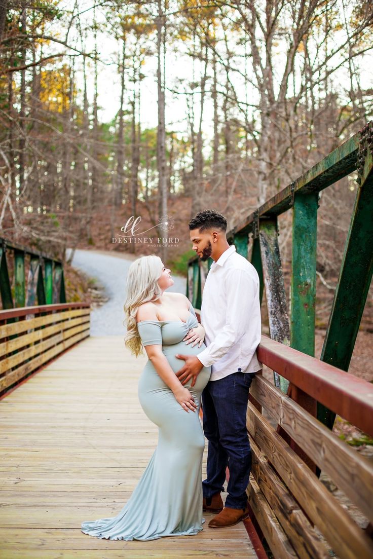 a pregnant woman standing next to a man on a bridge in front of trees and woods