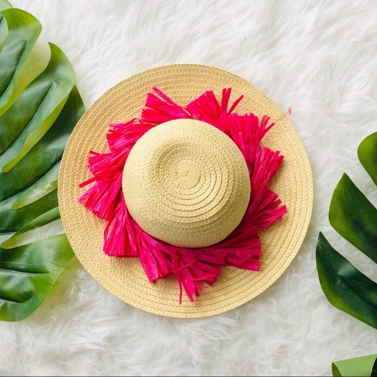 a straw hat with pink tassels sits on a white fur rug next to green leaves