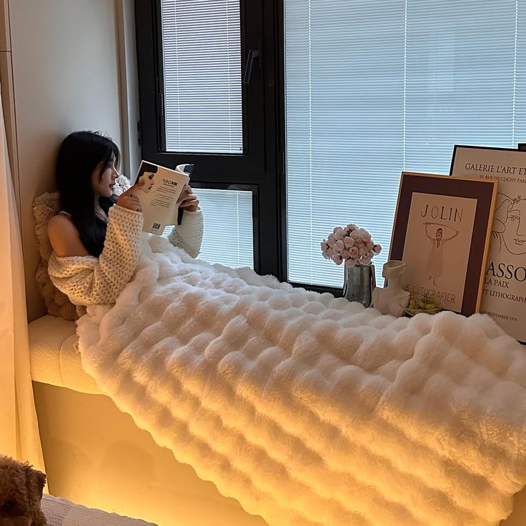 a doll sitting on a bed reading a book next to a window sill with books in it