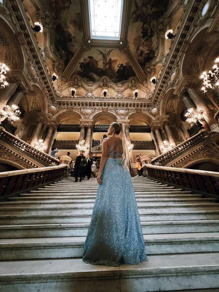 a woman in a blue dress is standing on some stairs and looking up at the ceiling