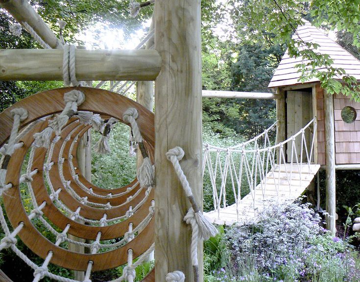 a rope bridge in the middle of a garden with trees and flowers around it, leading to a tree house