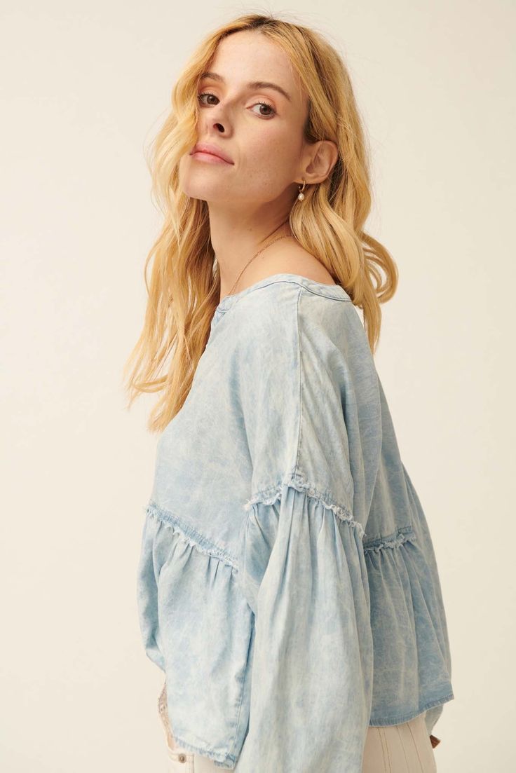 Acid washed denim blouse. Split neckline. 3-button closure with wood buttons. Long bubble sleeves with elastic cuffs. Drop shoulder. Exposed seams with frayed, raw-edge detailing. Flared ruffle hem. Babydoll silhouette. Loose fit. 100% Cotton. Imported. Designed in LA. Model wears size S. Fall Washed Relaxed Fit Blouse, Relaxed Fit Washed Blouse For Fall, Fall Washed Blue Chambray Denim Top, Fall Relaxed Fit Washed Blouse, Light Wash Chambray Denim Top, Relaxed Fit Medium Wash Denim Top, Spring Long Sleeve Washed Denim Top, Relaxed Fit Washed Tencel Tops, Fall Cotton Washed Blouse