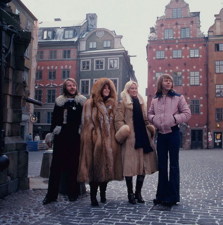 three women in fur coats stand on a cobblestone street with old buildings behind them