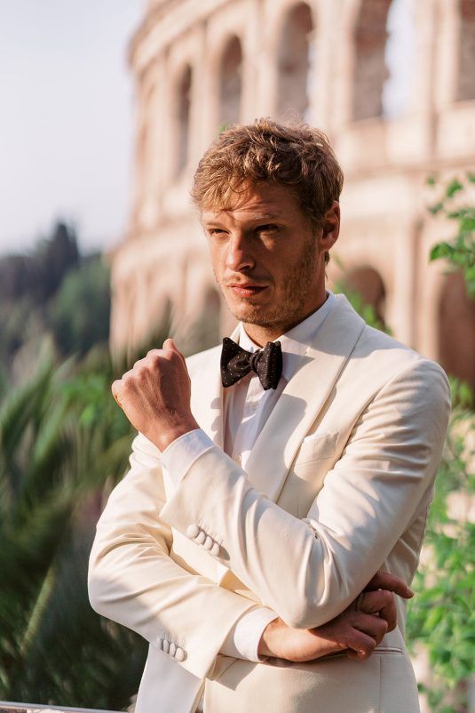 a man wearing a white suit and bow tie standing in front of an old building