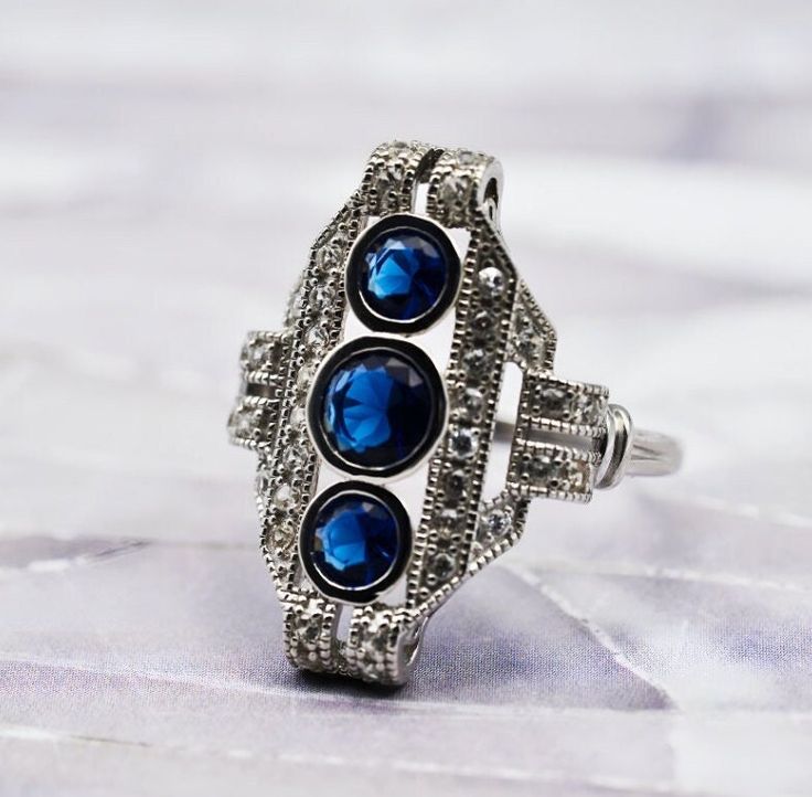 This exquisite vintage anti-tarnish rhodium plated ring is meticulously crafted from 925 solid sterling silver. It is adorned with three sapphire stones as its centerpiece, which are complemented by sparkling cubic zirconia accent stones. The dimensions of the ring are notable, with the sapphire arrangement measuring 0.90 inches in length and 0.65 inches in width, creating a striking and elegant presence on the finger. This piece is a testament to classic style and timeless craftsmanship. Stampe Classic Silver Crystal Ring With Birthstone, Elegant Silver Crystal Ring With Birthstone, Elegant Silver Crystal Birthstone Ring, Vintage Sapphire Jewelry For Anniversary, Vintage Cubic Zirconia Gemstone Jewelry, Vintage Diamond White Sapphire Jewelry, Art Deco Ring With Accent Stones For Gifts, Art Deco Rings With Accent Stones For Gifts, Antique Cubic Zirconia Jewelry Gift