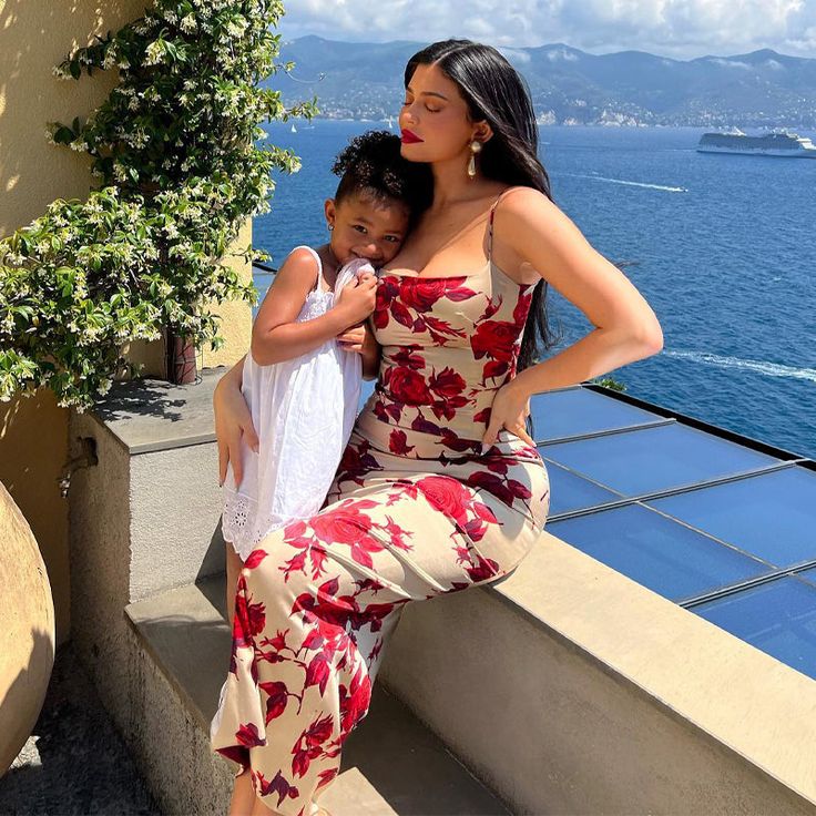 a woman in a floral print dress poses with a young child on a balcony overlooking the ocean