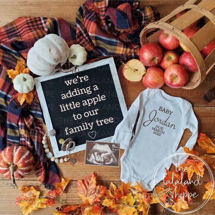 baby clothes, apples and pumpkins are laid out on a wooden table with the words we're adding a little apple to our family tree