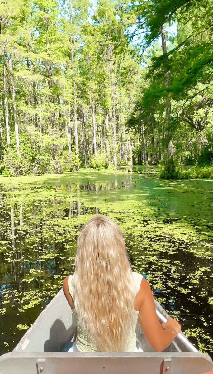 a woman riding in a boat down a river surrounded by green trees and water lillies