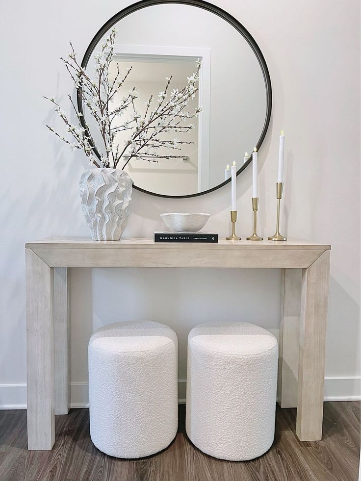 two white stools under a round mirror on the wall next to a table with candles