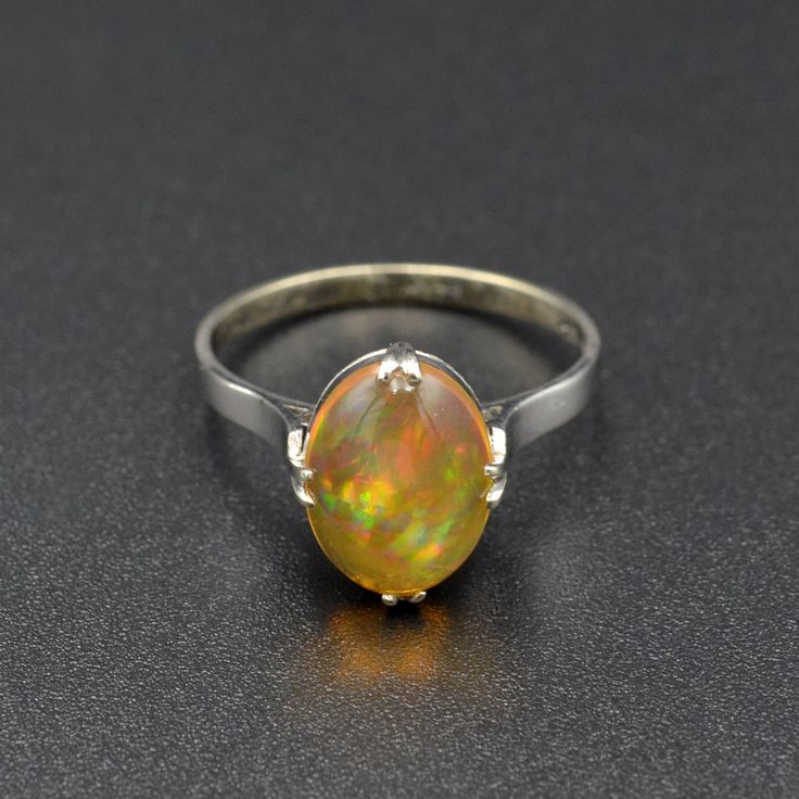 "A rainbow is swimming in this gorgeous antique opal ring! A beautifully proportioned opal cabochon is centered with cardinal prongs (north, south, west and east) in an 18k white gold setting. The opal features a transparent orange body, typical of Mexican jelly opals, with a \"contra luz\" color flash seen when the gem interacts with light. The color play is astounding, with every color of the rainbow ignited within the body of the opal, having painterly brush-stroke like patterns. This ring hails from the 1920s and has been tenderly well-cared for in its lifetime. It is in wonderful condition and ready for a new owner. This ring could be a perfect alternative engagement ring, gift for someone with an October birthstone or simply a present for an antique jewelry lover.   Opal is a fascina Classic Opal Moonstone Ring As Gift, Vintage Opal Ring As A Gift, Oval Cabochon Opal Ring With Polished Finish, Vintage Opal Ring With Polished Finish For Formal Occasions, Classic Oval Opal Ring With Polished Finish, Vintage Oval Cabochon Gemstones, Fine Jewelry Oval Cabochon Opal Ring, Ethiopian Opal Oval Rings For Anniversary, Oval Cabochon Opal Ring Fine Jewelry