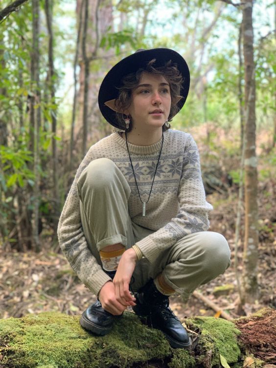 a young man sitting on top of a moss covered rock in the woods wearing a hat