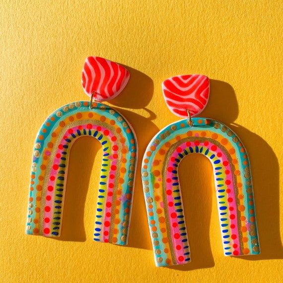 two pairs of colorful painted wooden earrings on a yellow background, one with a rainbow design and the other with a red heart