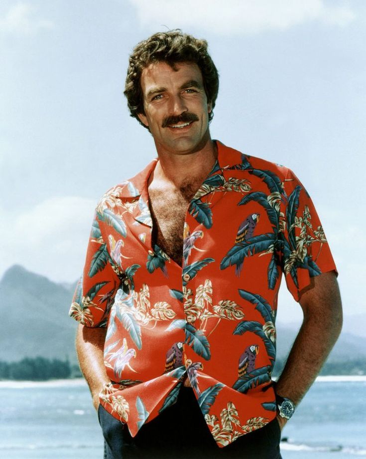 a man with a moustache standing in front of the ocean wearing a hawaiian shirt