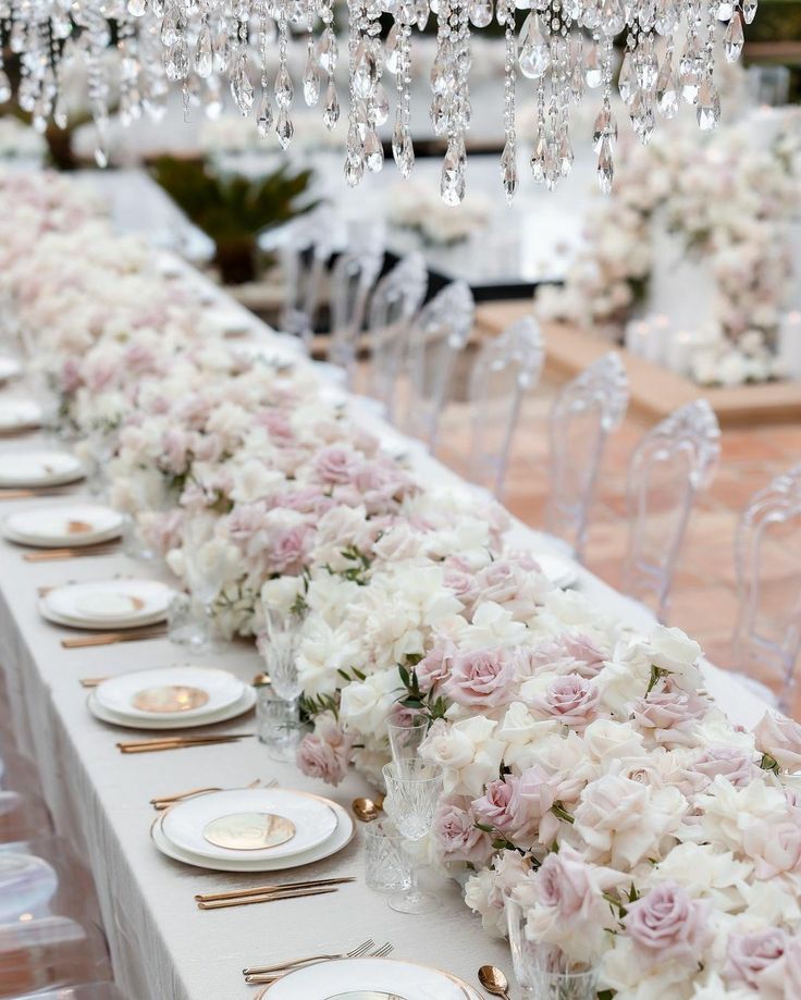 a long table is set with white and pink flowers, gold place settings and crystal chandeliers