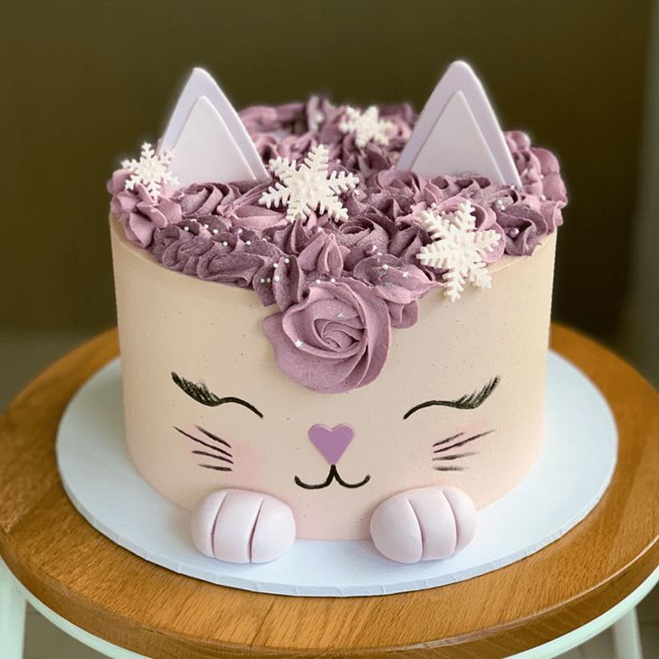 a cat cake with pink frosting and purple flowers on top, sitting on a table