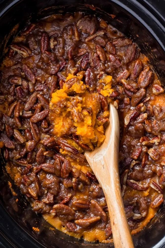 a wooden spoon in a slow cooker filled with sweet potato casserole and pecans