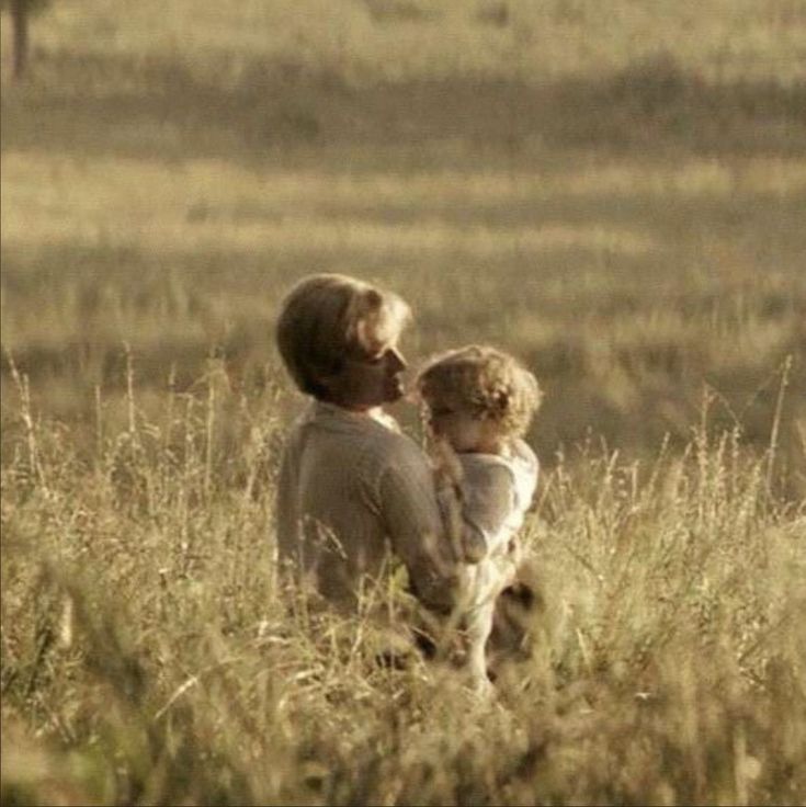 a woman holding a child in a field with tall grass and an elephant behind her