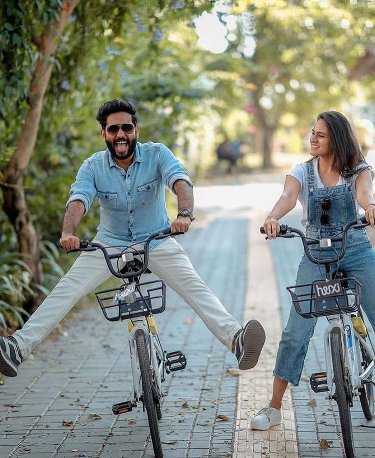 two people are riding bikes on the sidewalk with trees in the background and one person is smiling at the camera