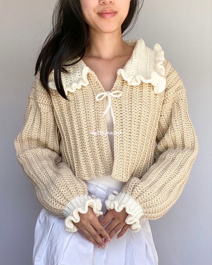 a woman wearing a sweater with ruffles on the collar and sleeves, standing in front of a gray wall