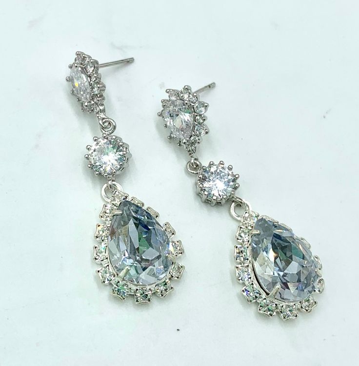 "* Crowned with brilliant CZ Teardrop Earrings, (available in Posts or Clip-Ons,) are CZ Connectors and gorgeous 14mm Swarovski® Dusty Blue Teardrop Pendants. Each Dusty Blue Shade Teardrop is surrounded by many Swarovski Clear Rhinestones. You may also select a different color Swarovski stone if you wish * The total length of each dangling Wedding Earring is 1 3/4\" (46mm). * Settings are cast in brass and are overlaid with STERLING SILVER. Rose Gold or Gold Overlay Accompanying the beautiful W Dazzling Crystal Teardrop Bridal Earrings, Silver Crystal Drop Bridal Earrings, Diamond White Teardrop Crystal Bridal Earrings, Silver Teardrop Bridal Earrings With Sparkling Stones, Silver Teardrop Crystal Bridal Earrings, Dazzling Sparkling Drop Bridal Earrings, Dazzling Drop Bridal Earrings With Sparkling Stones, Silver Teardrop Pendant Earrings For Wedding, Diamond White Bridal Earrings With Sparkling Stones