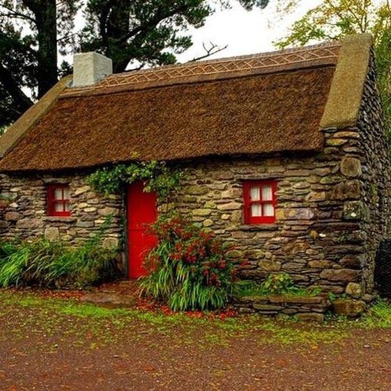 an old stone house with a red door and thatched roof is surrounded by greenery