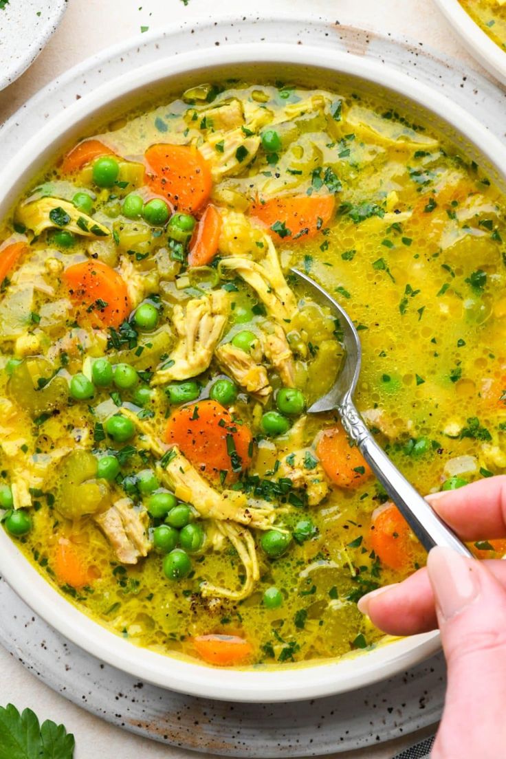 a person holding a spoon over a bowl of chicken and veggie soup with carrots