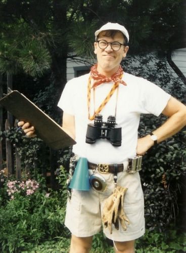 a man in white shirt and shorts standing next to trees with binoculars on his chest