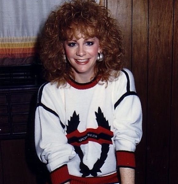 a woman standing in front of a wooden wall wearing a white sweater with black and red designs on it