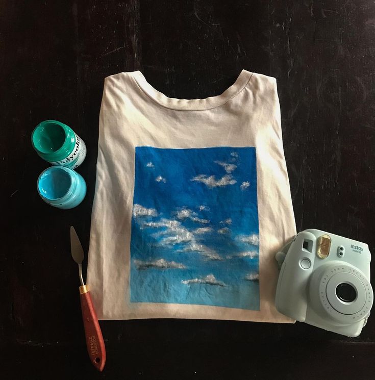 a white t - shirt with blue sky and clouds on it next to a pair of scissors