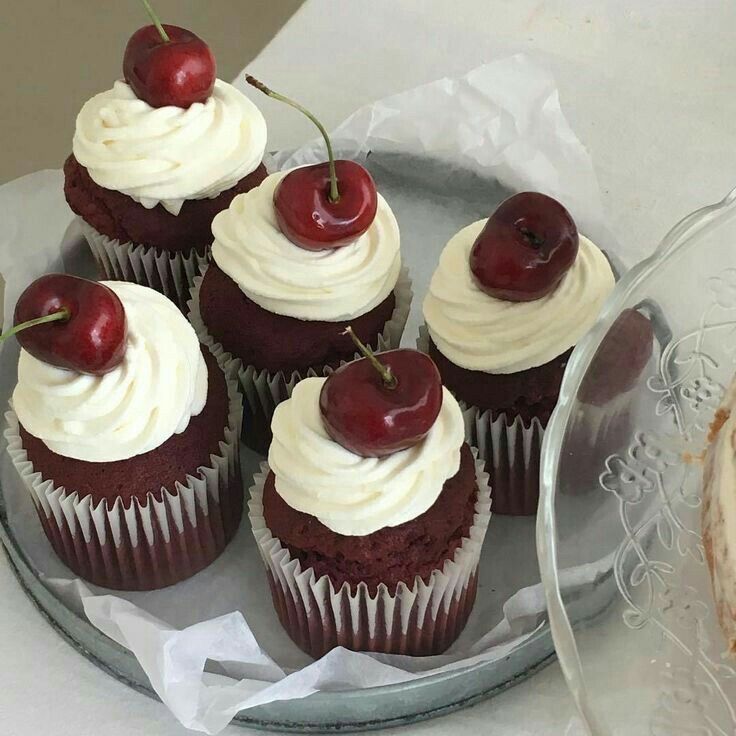 cupcakes with white frosting and cherries are on a glass platter