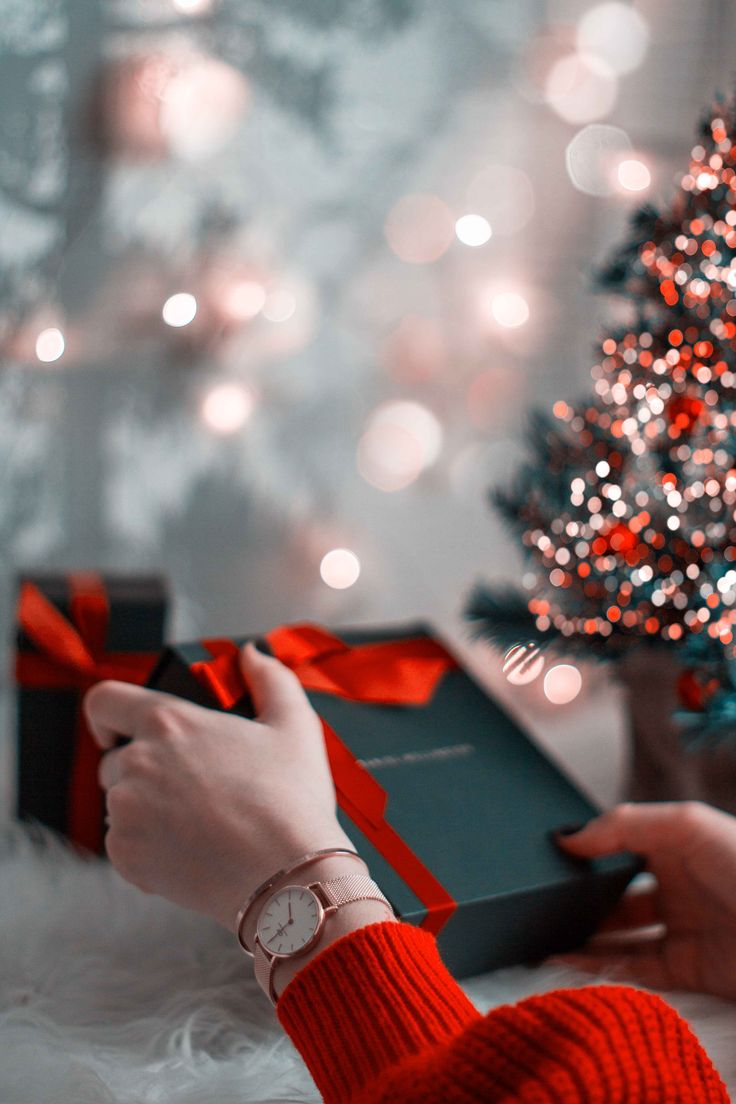 a woman is opening a gift box in front of a christmas tree with red and white lights