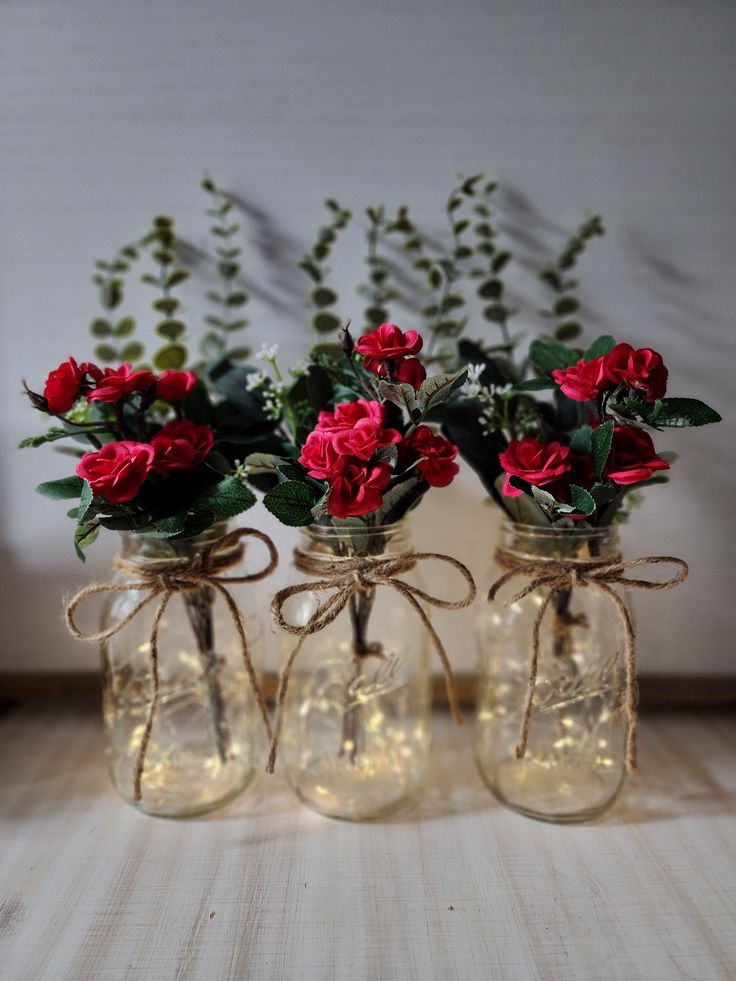 three mason jars with red roses tied in twine and some greenery are sitting on a table