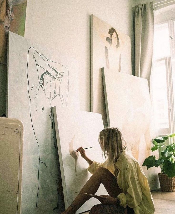 a woman sitting on the floor in front of some paintings and holding a paintbrush