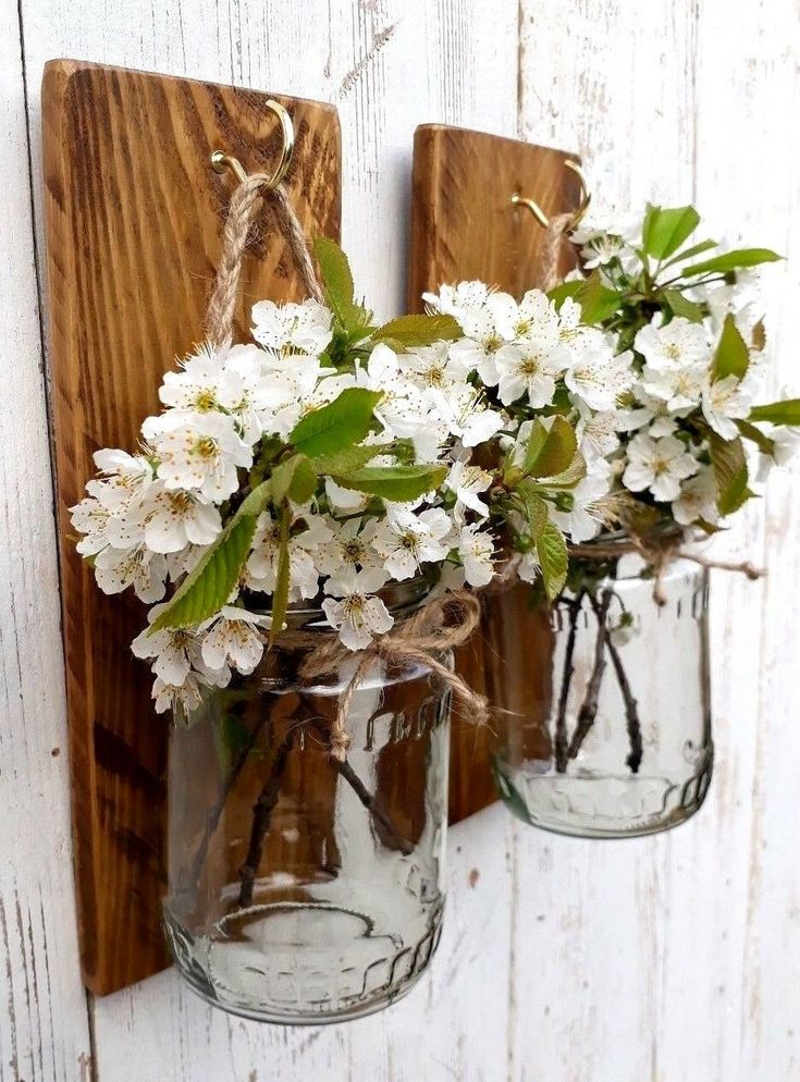 two mason jars with white flowers are hanging on a wooden wall next to each other