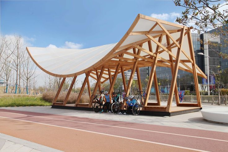 a group of people sitting under a wooden structure on the side of a road next to a building