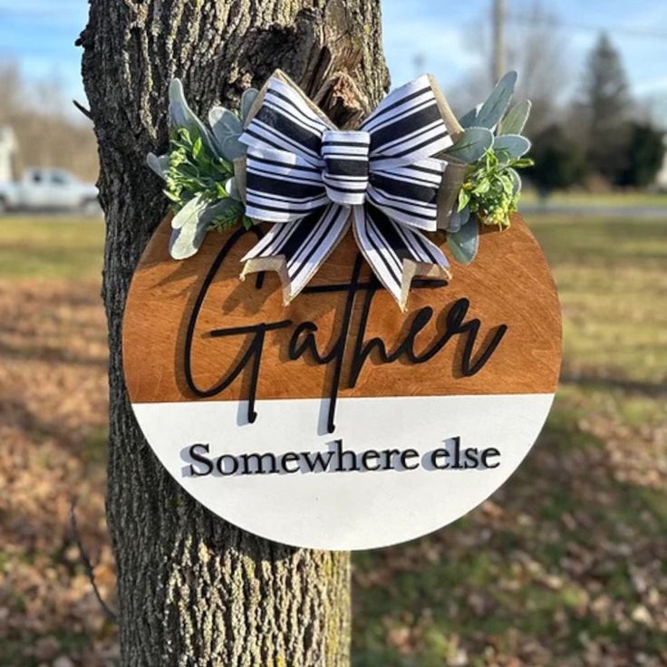 a wooden sign that says gather somewhere else hanging on a tree in front of a field