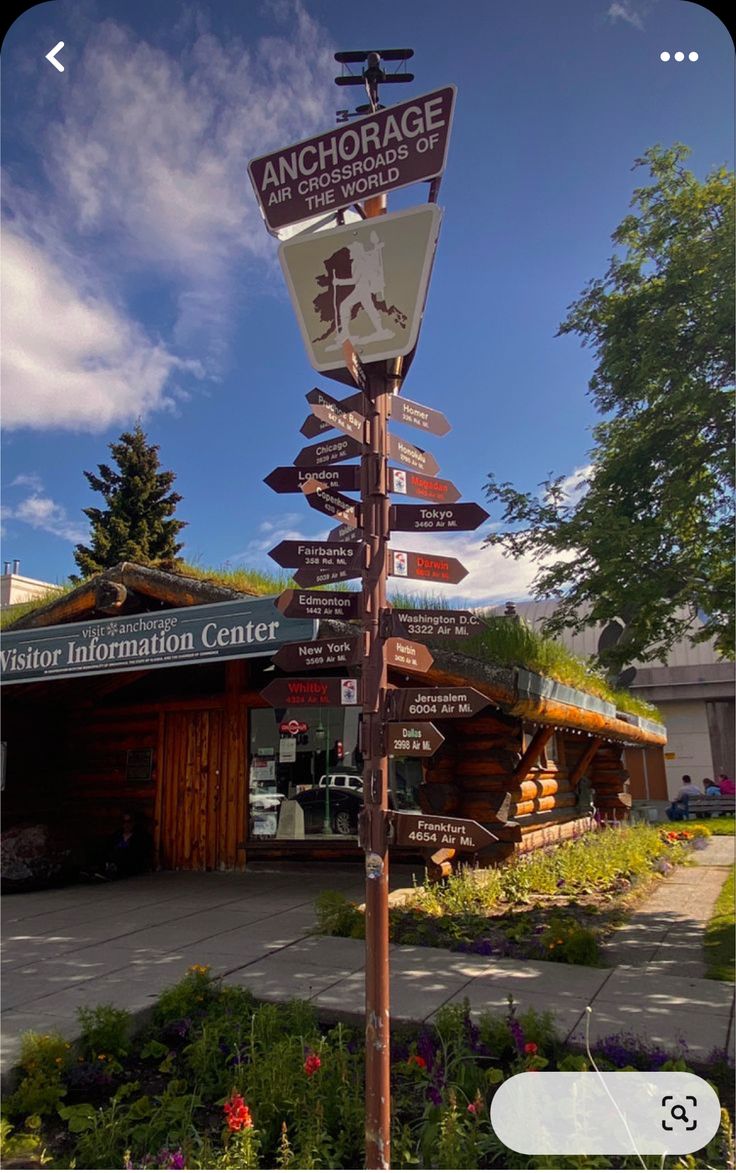 a pole with many signs on it in front of a building and some flowers outside