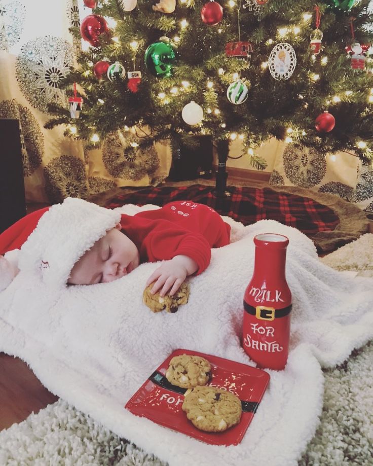 a baby laying on a blanket next to a christmas tree with cookies and a bottle of hot sauce