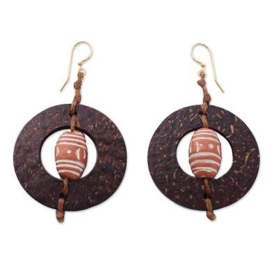 Ornate terracotta beads center coconut shells in the design of these bold earrings. The design of the earrings makes me think of medieval architecture says Nonyem Abena Ibeneme. The earrings are crafted by hand with waxed linen cords and brass hooks. Artisan Brown Beaded Earrings For Pierced Ears, Bohemian Brown Pierced Hoop Earrings, Handmade Bohemian Brown Plug Earrings, Artisan Brown Earrings For Pierced Ears, Handmade Brown Bohemian Plug Earrings, Fair Trade Brown Jewelry With Round Beads, Brown Beaded Drop Earrings For Beach, Brown Fair Trade Round Bead Jewelry, Brown Round Bead Earrings For Festivals