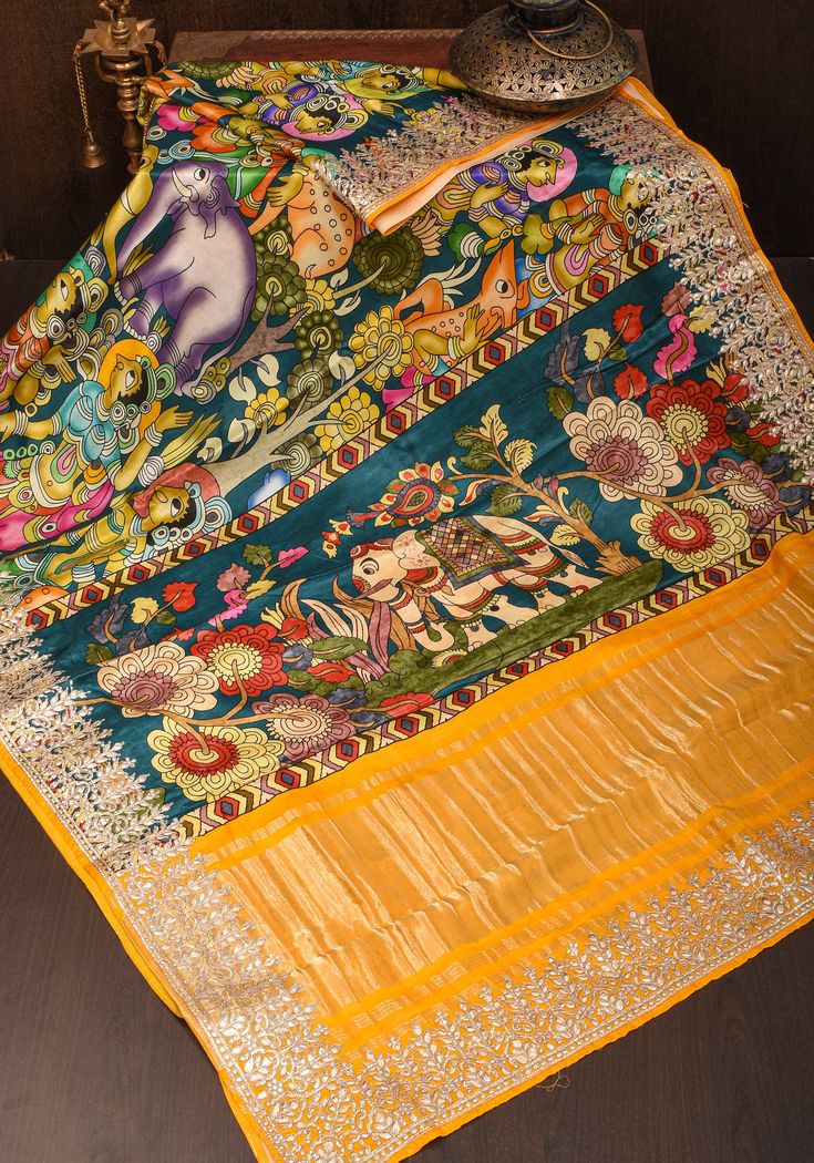 A significant style of embellishing special occasion clothes, the Gotta Patti technique from Rajasthan adds shimmer and highlight! It involves an applique technique with metallic trims in unique designs that are sewn to the garment. Initially crafted for the Royals, this technique has proudly stood the test of time and remains coveted! This Gajji Silk Saree is soft and lustrous and comes in a vivid Kalamkari digital print. The gotta patti border and crushed zari panel enhance its look and makes it stand out.  The saree is ready to wear with falls and pico done, an unstitched blouse fabric is included. Note: Colors that show up on your screen may vary slightly from the actual product due to variations in settings. Also, the actual product may appear to be different hues depending on the amb Navratri Multicolor Pre-draped Saree With Motifs, Chanderi Meenakari Pre-draped Saree For Navratri, Festive Multicolor Pre-draped Saree With Motifs, Gold Embroidered Silk Pre-draped Saree, Festive Multicolor Chanderi Pre-draped Saree, Multicolor Pre-draped Saree With Zari Work For Puja, Festive Multicolor Unstitched Pre-draped Saree, Multicolor Chanderi Pre-draped Saree With Zari Work, Multicolor Tussar Silk Pre-draped Saree For Eid