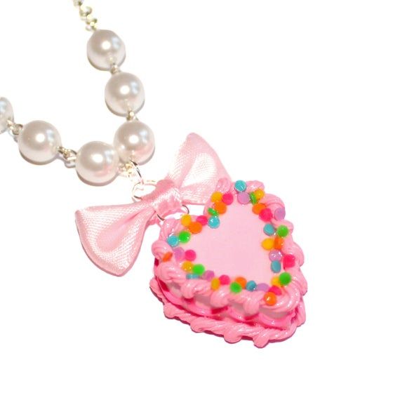 This adorable miniature birthday cake necklace is a special way to celebrate your birthday or make everyday your birthday! It was inspired by all those fabulous kitschy and girly pink birthday cakes we had growing up.This cake is adorned with classic birthday cake details like pink piped frosting, frilly bright pink icing along the middle, and topped with bright rainbow confetti sprinkles. The cake hangs below a lovely pink bow from a high quality glass pearl beaded chain and silver plated chain Pink Heart-shaped Charm Necklace For Party, Pink Heart Charm Necklace For Party, Sweet Pink Necklace For Valentine's Day, Sweet Pink Necklaces For Valentine's Day, Charming Pink Party Jewelry, Pink Kawaii Jewelry For Valentine's Day, Cute Valentine's Day Party Necklaces, Kawaii Pink Jewelry For Valentine's Day, Personalized Kawaii Jewelry For Birthday