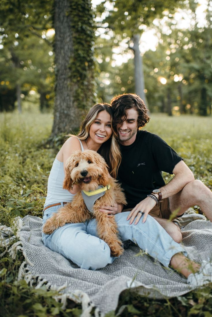 a man and woman sitting on a blanket with a dog in their lap smiling at the camera