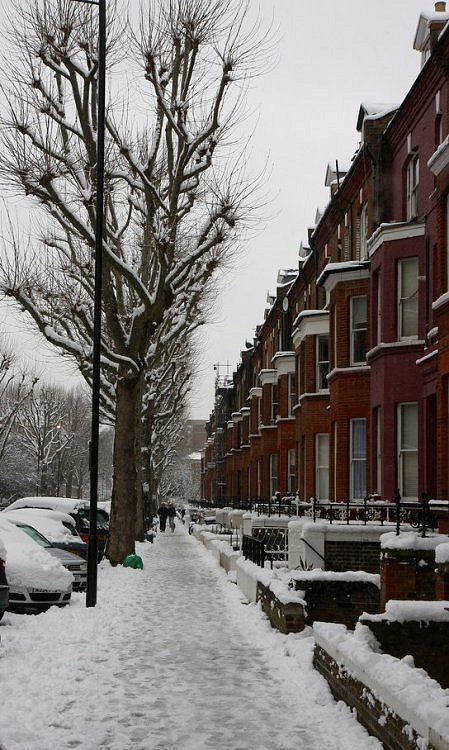 a snow covered street with cars parked on the side and people walking down the sidewalk