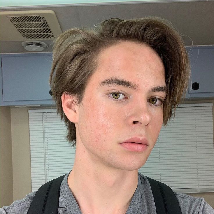 a young man with brown hair and grey shirt looking at the camera while wearing a black backpack